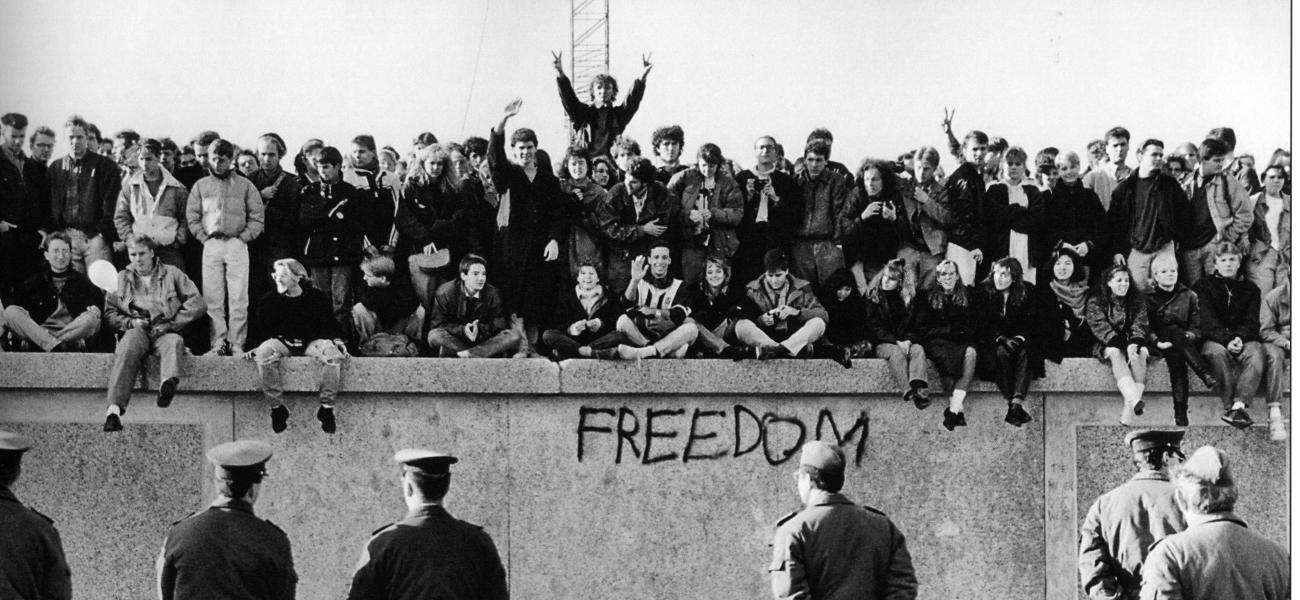 East German students sit atop the Berlin Wall at the Brandenburg Gate in front of border guards, November 1989.
